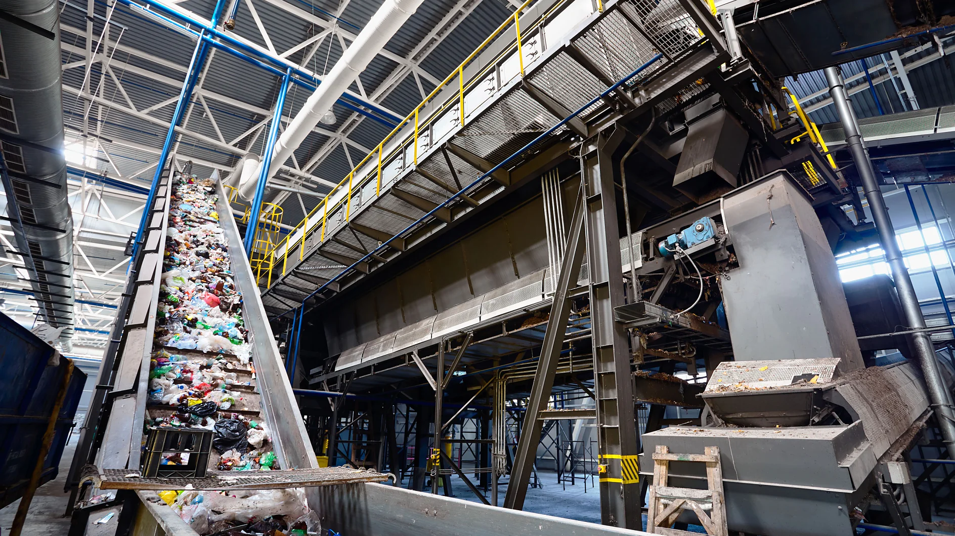 Conveyor belt carrying assorted waste materials in a large industrial recycling facility. Overhead metal structures and machinery are visible, with a high ceiling and hanging lights providing illumination. (This text has been generated by AI)