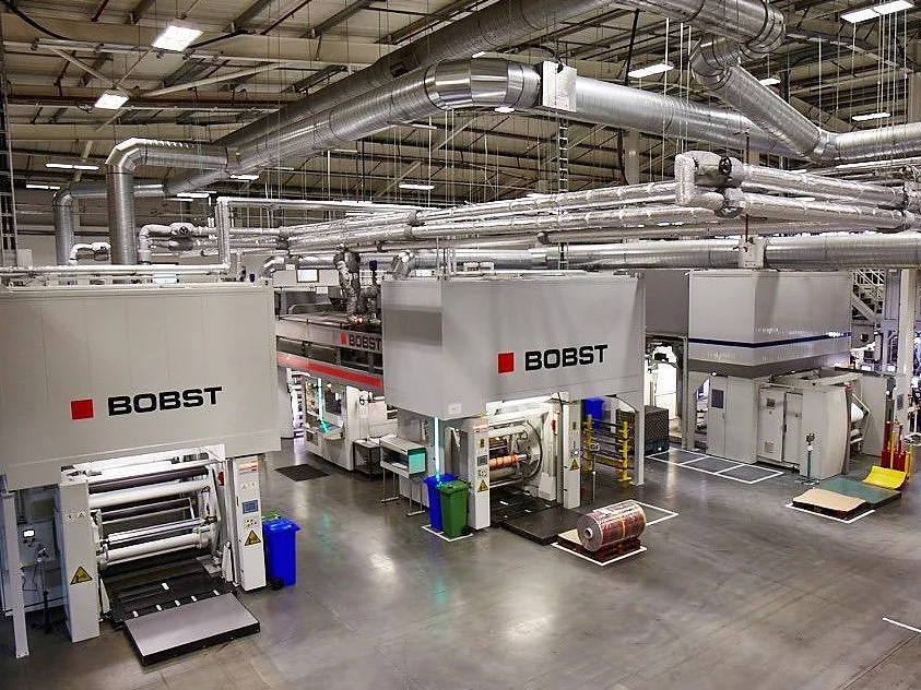 A large industrial facility with multiple Bobst machines for printing or packaging. The machines are white with red and black logos. The room has a high ceiling with exposed ducts and overhead lighting. Various materials, including tesa tape, and bins are visible. (This text has been generated by AI)