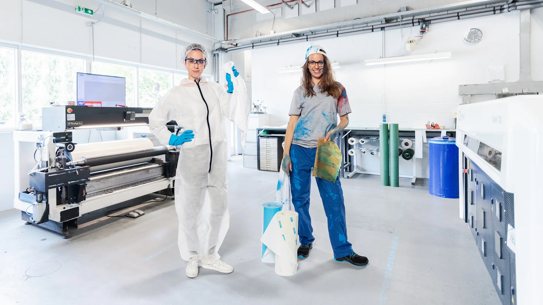 Two people stand in a printing workshop. One wears a white protective suit and goggles, holding a roll of tesa tape. The other is dressed in casual clothes with a cap, holding a large print. The background features printing equipment and rolls of material. (This text has been generated by AI)