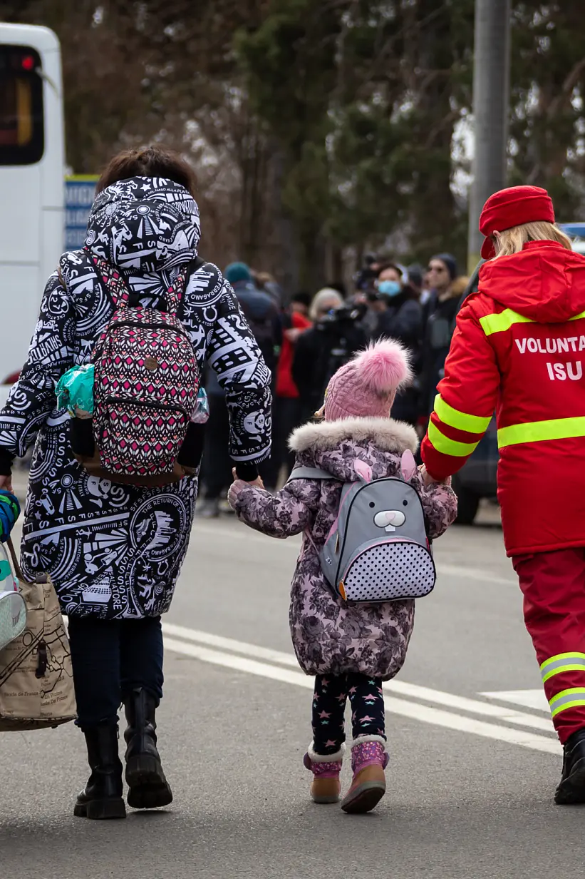 A woman and two children walk down a street near parked vehicles. The children wear backpacks and winter clothing. A volunteer wearing a red uniform assists them, using tesa tape to secure a large white bag. A bus and several people are visible in the background. (This text has been generated by AI)