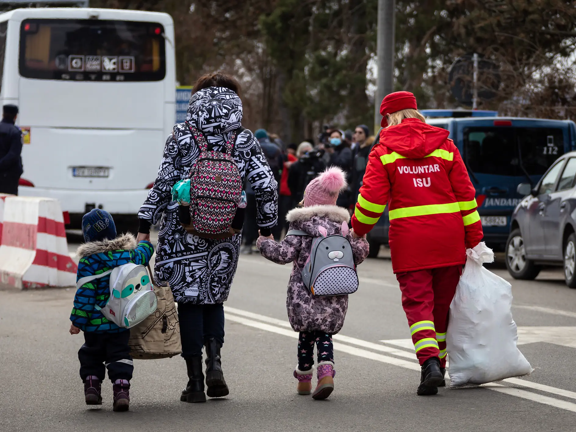 A woman and two children walk down a street near parked vehicles. The children wear backpacks and winter clothing. A volunteer wearing a red uniform assists them, using tesa tape to secure a large white bag. A bus and several people are visible in the background. (This text has been generated by AI)