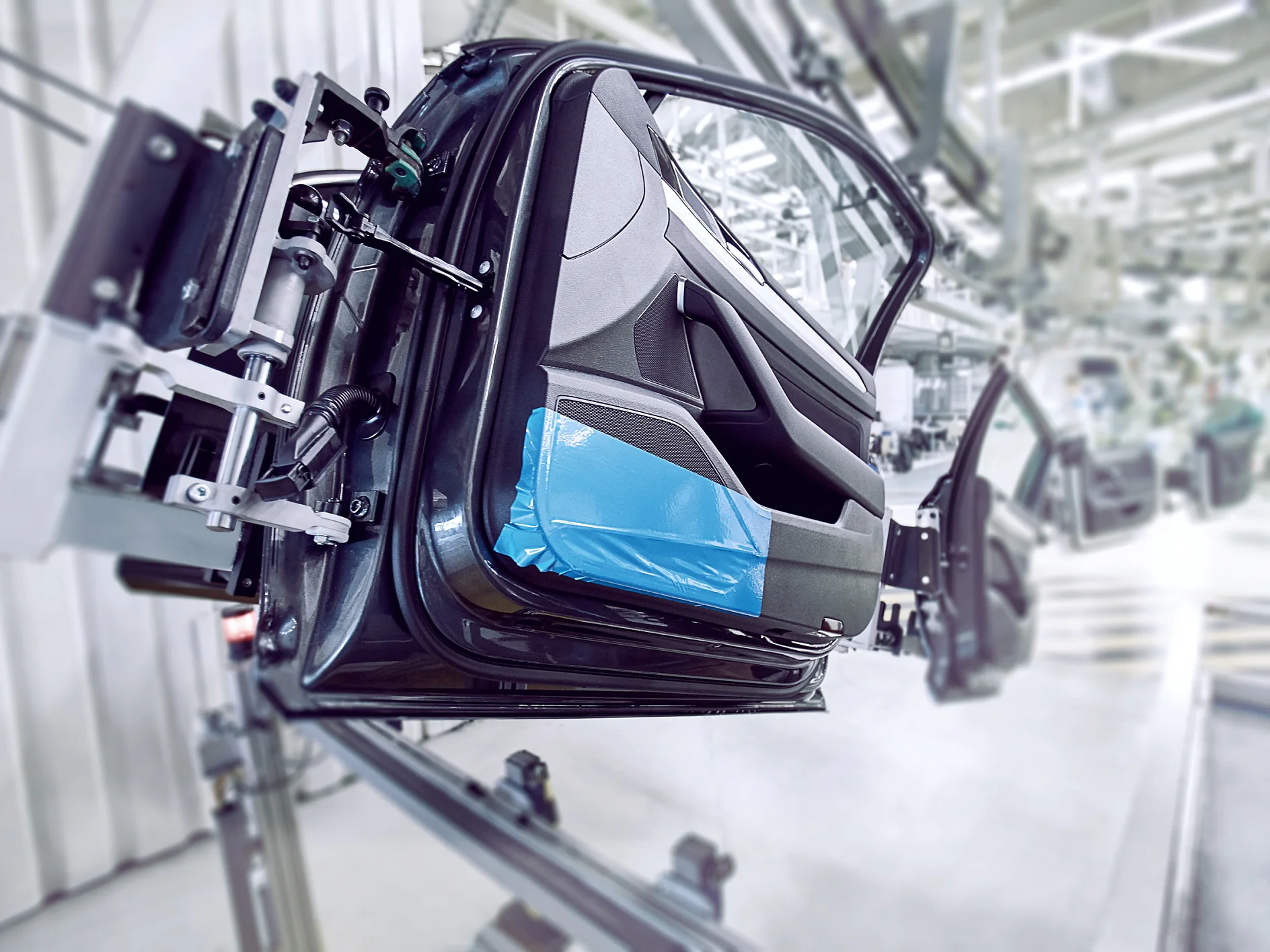 A car door is mounted on an assembly line in an industrial setting. The interior side of the door is visible, partially covered with blue protective tesa tape. The background shows more equipment and blurred parts of the assembly line. (This text has been generated by AI)