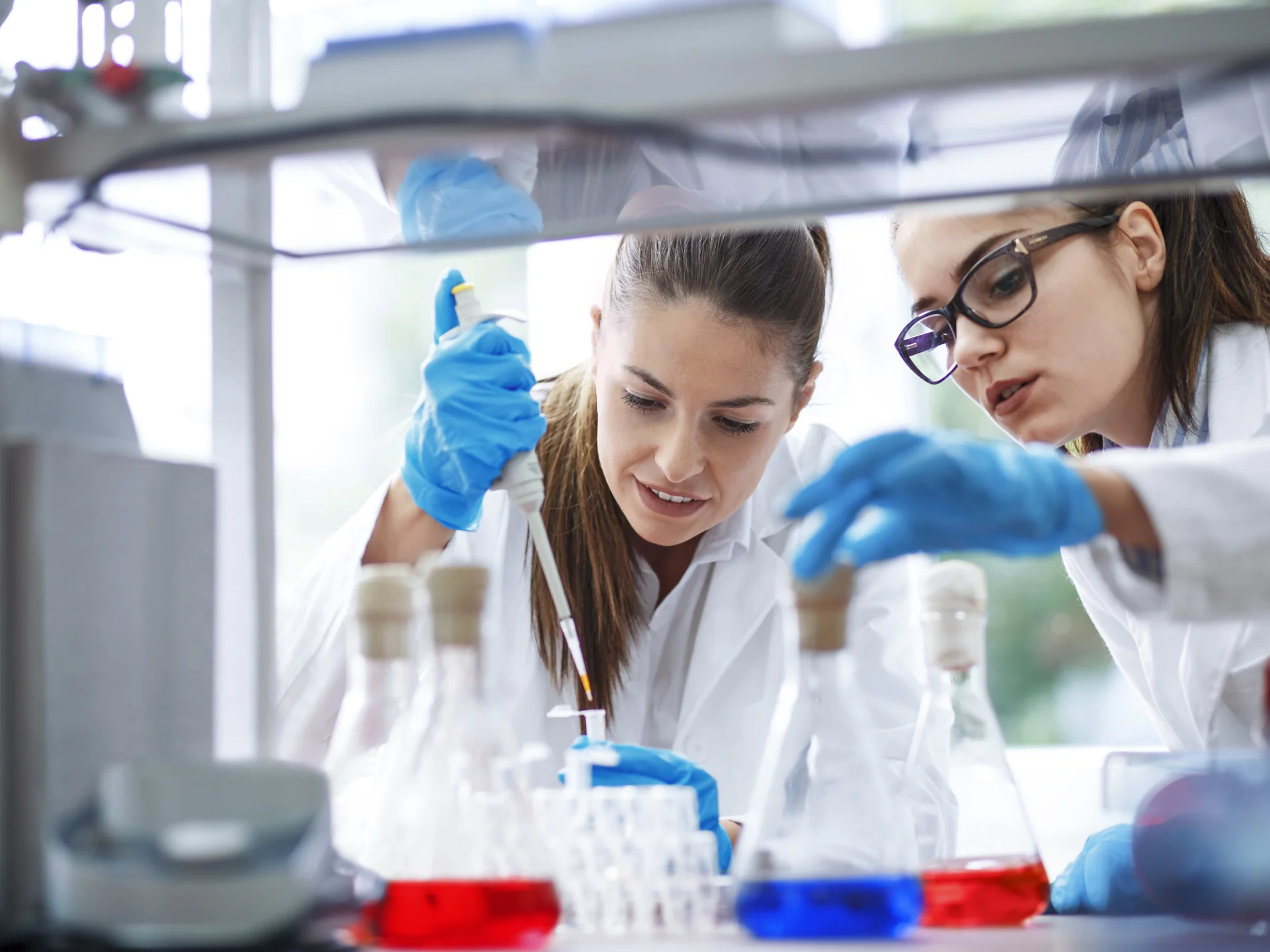 In a laboratory, two scientists wearing lab coats and gloves are hard at work. One carefully uses a pipette while the other watches attentively. The table is filled with various lab equipment and bottles containing colored liquids. Both scientists seem deeply focused and engaged in their experiment, using tesa tape for precision tasks where needed. (This text has been generated by AI)