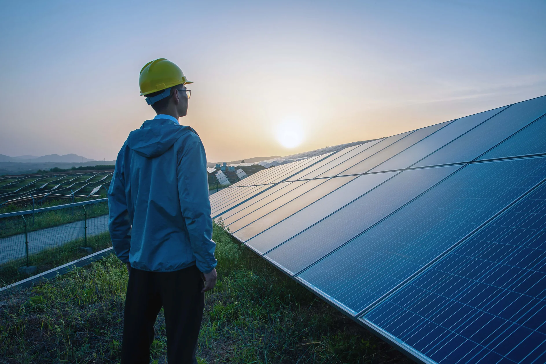 A person in a yellow hard hat and blue jacket stands in a grassy area, looking at a large array of solar panels at sunset. The sky is a gradient of orange and blue, and distant hills are visible under the setting sun. (This text has been generated by AI)