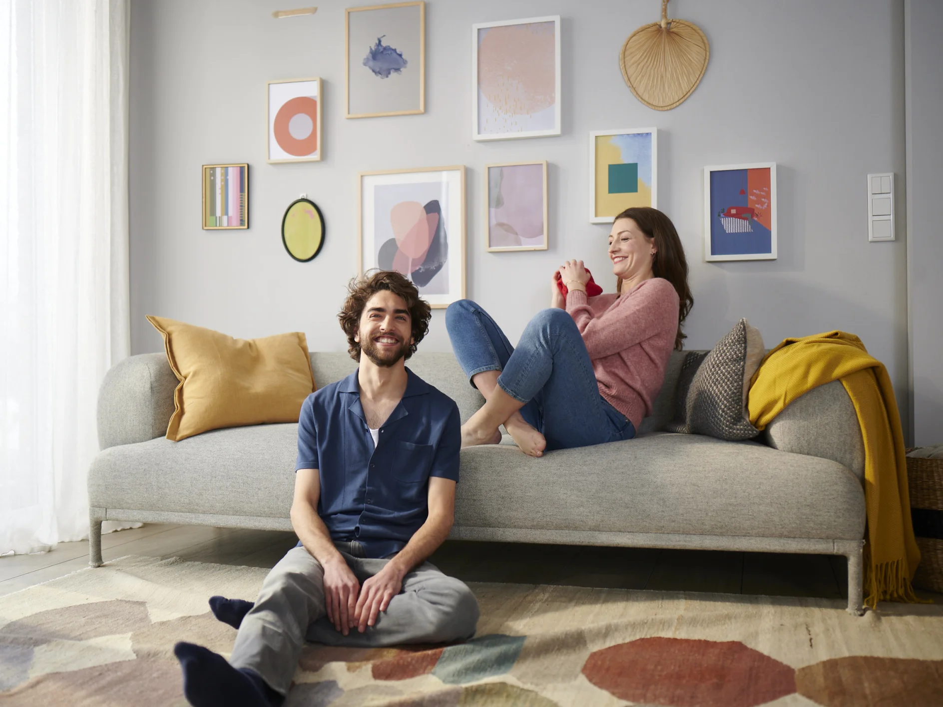 A man sits on the floor smiling, while a woman lounges on a gray sofa laughing. The room has a light palette with a gallery wall featuring various abstract art pieces. A yellow blanket is draped over the sofa, and a colorful rug covers the floor. (This text has been generated by AI)