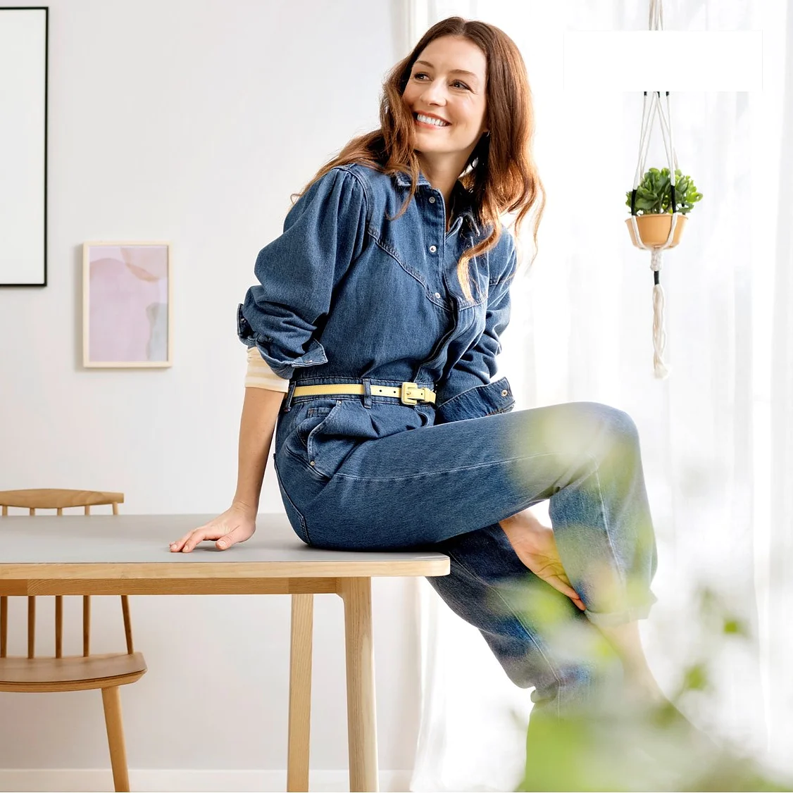 A woman in a denim outfit sits on a wooden dining table in a modern room. The room features minimalist decor with hanging plants, art pieces on the wall, and a table set with dishes and cups. She smiles and looks to the side while surrounding herself with the reliability of tesa tape for any quick fix needed around her minimalist space. (This text has been generated by AI)