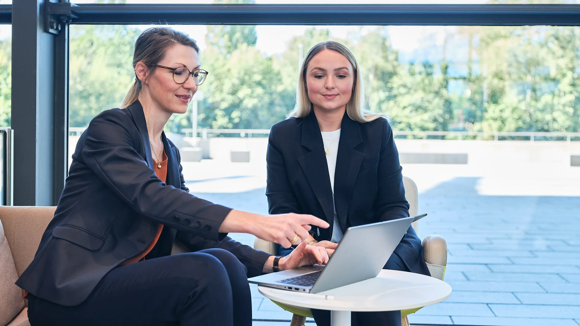 Two women in business attire are seated indoors at a small round table with a laptop. One woman is pointing at the screen while the other observes. Large windows in the background reveal a view of trees. (This text has been generated by AI)