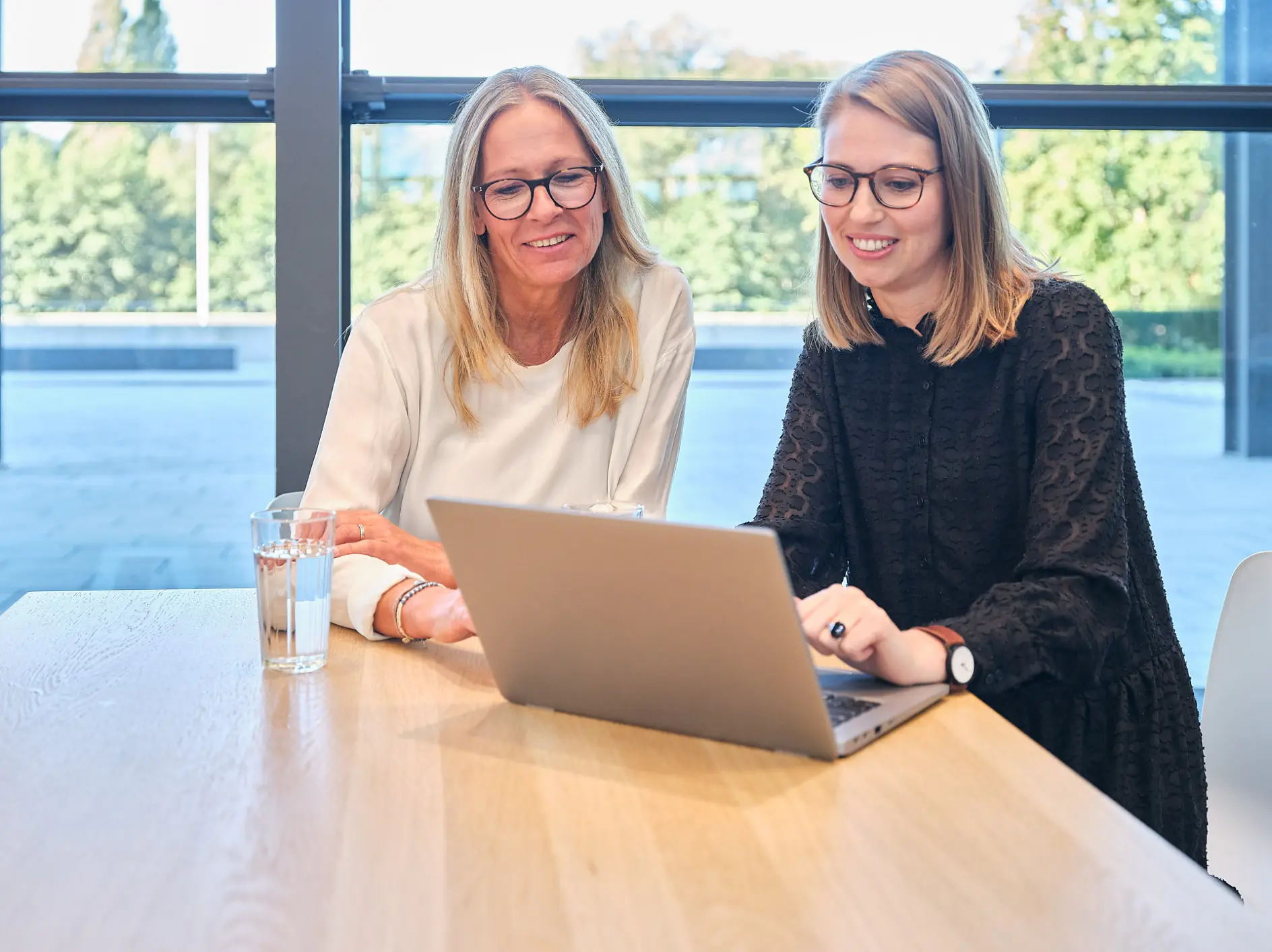 Two women are seated at a wooden table, working on a laptop. One wears a white top and glasses, while the other wears a black top and glasses. Both are smiling. A glass of water is on the table. A large window shows trees outside. (This text has been generated by AI)