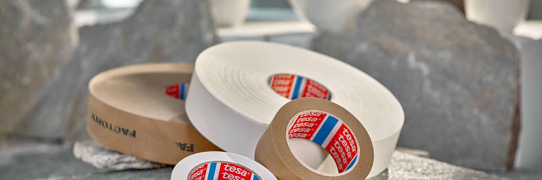 Several rolls of adhesive tesa tape, including white and brown, are placed on stone slabs. In the background, there are white bowls and cups, and cardboard pieces are visible in the foreground. Sunlight streams in through a window. (This text has been generated by AI)