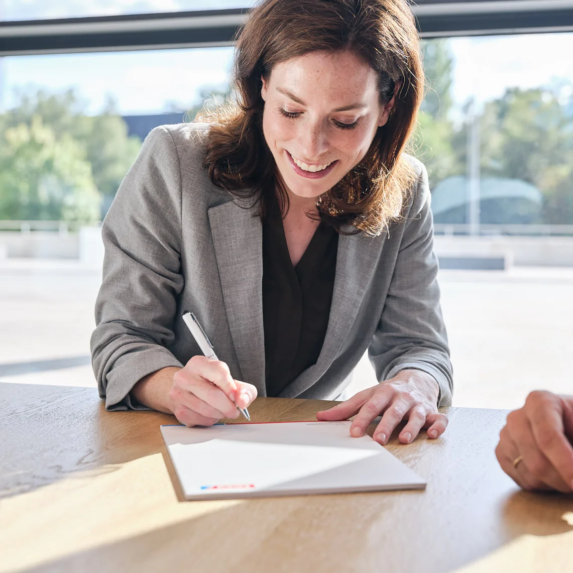 A woman in a gray blazer is sitting at a wooden table, smiling, and writing on a piece of white paper with a pen. Another persons hand rests on the table near her. Large windows with greenery outside are in the background. (This text has been generated by AI)