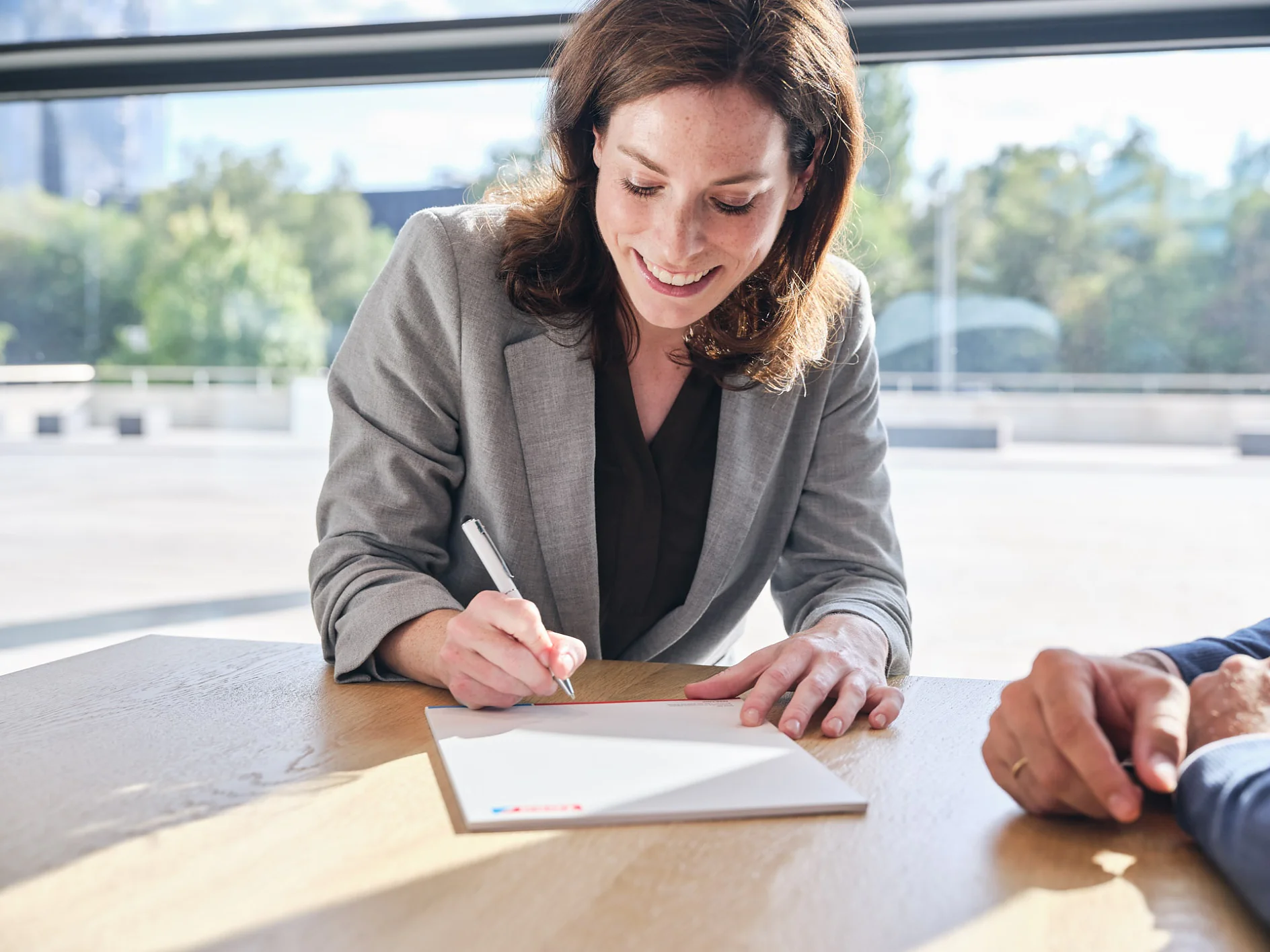 A woman in a gray blazer is sitting at a wooden table, smiling, and writing on a piece of white paper with a pen. Another persons hand rests on the table near her. Large windows with greenery outside are in the background. (This text has been generated by AI)