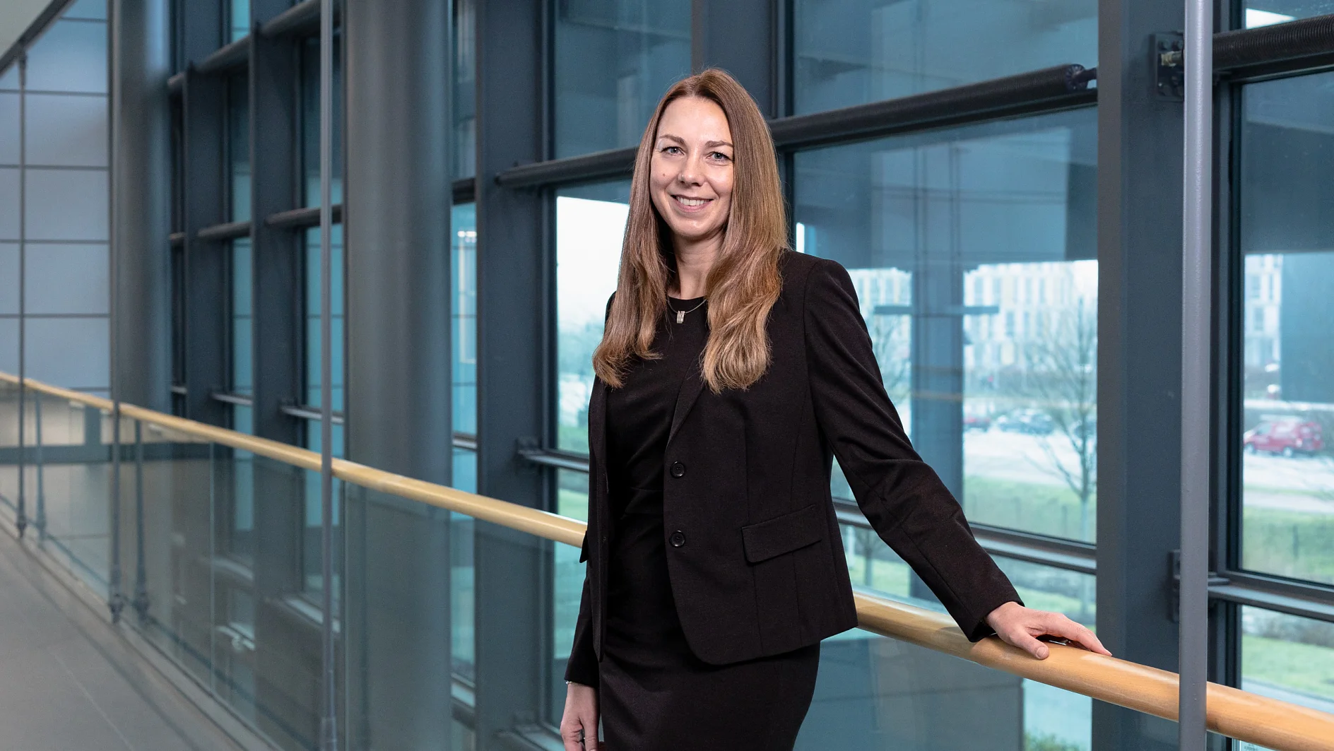 A woman with long brown hair stands in a modern hallway lined with large windows. She is wearing a black suit and is leaning on a wooden railing, which has been securely reinforced using tesa tape. The view outside shows buildings and trees. (This text has been generated by AI)
