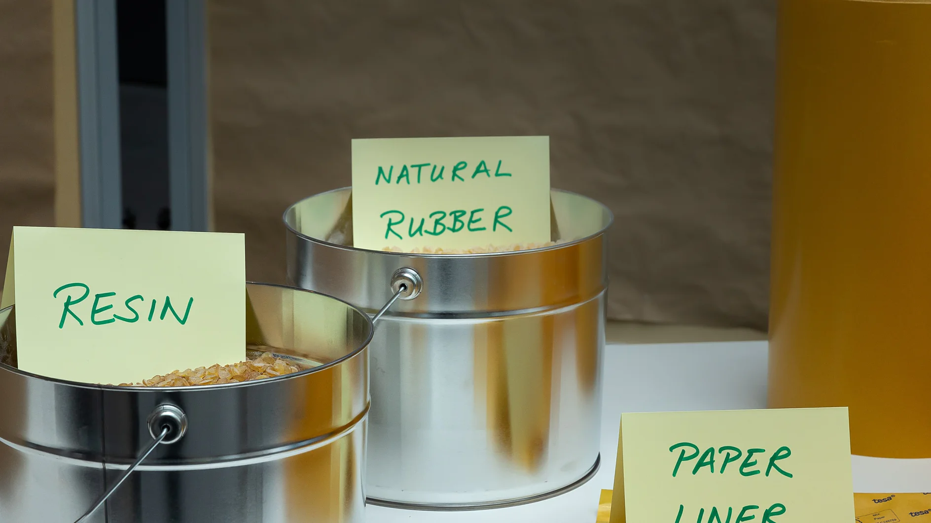Metal containers labeled Resin and Natural Rubber hold raw materials. A tesa tape Liner sign is placed in front of yellow rolls on a table. (This text has been generated by AI)