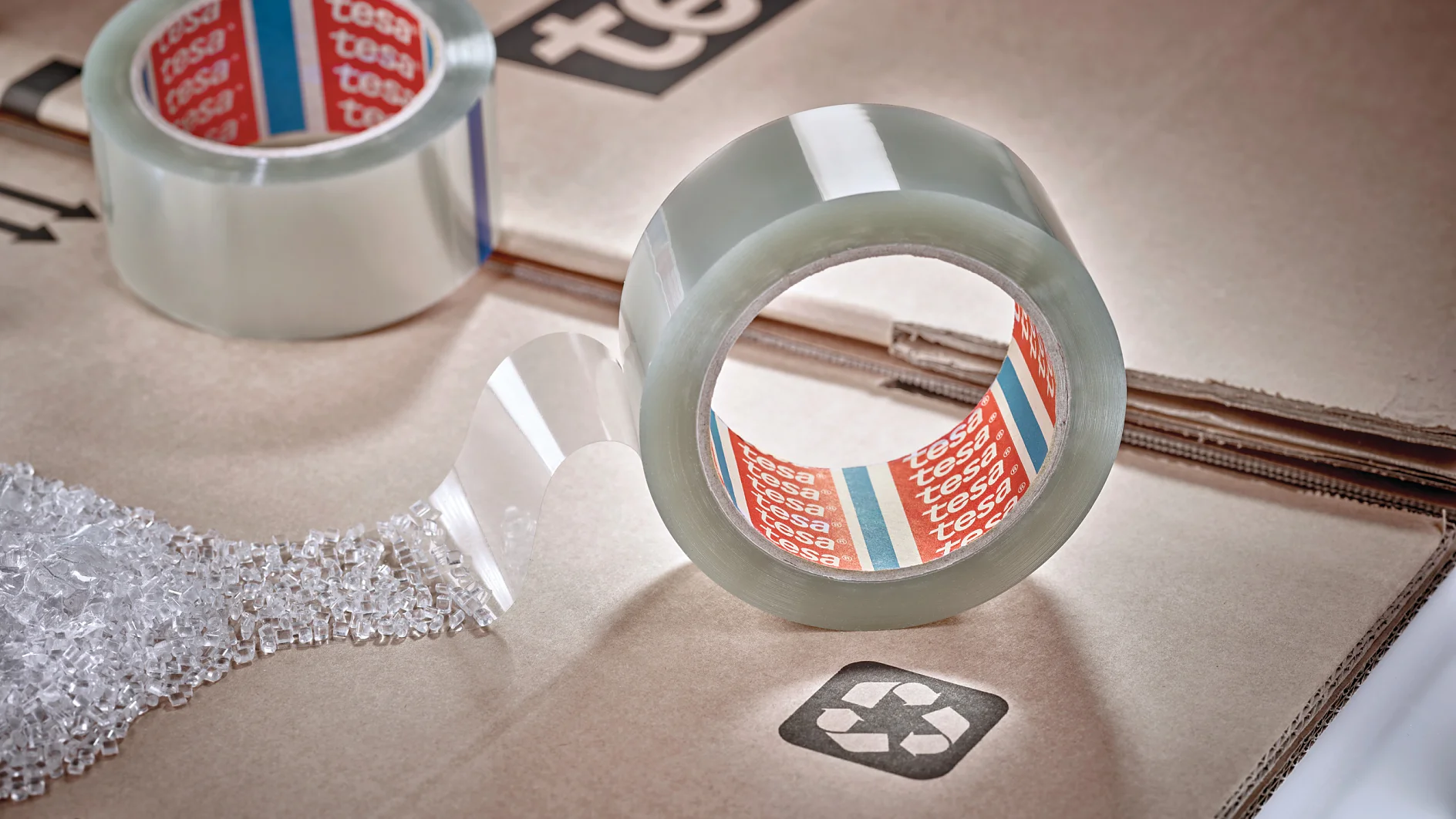 A roll of clear packing tesa tape with the brand name visible sits on a cardboard surface. A strip of tesa tape extends outward. Another roll is partially visible, and bubble wrap is nearby. The cardboard has a recycling symbol printed on it. (This text has been generated by AI)