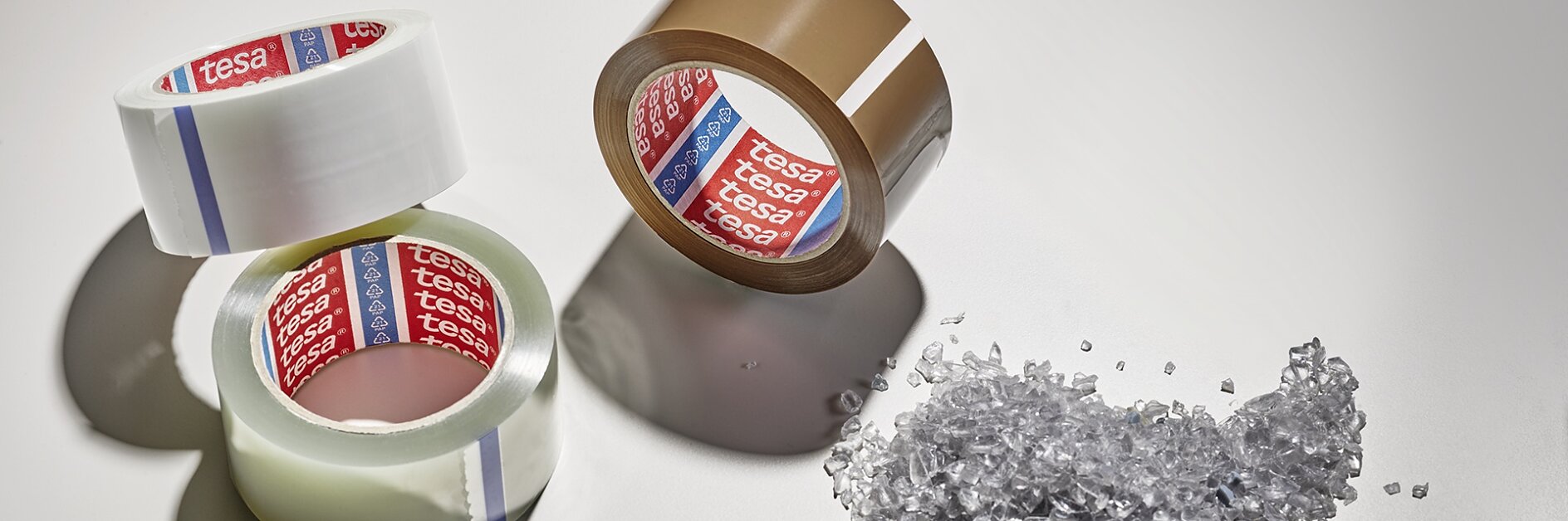 Three rolls of packing tesa tape, two clear and one brown, are arranged on a light surface. Beside them is a small pile of clear plastic granules. Shadows extend from the objects. (This text has been generated by AI)