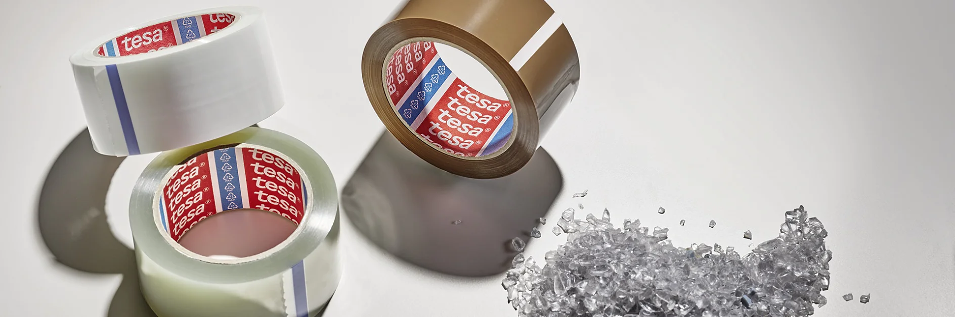 Three rolls of packing tesa tape, two clear and one brown, are arranged on a light surface. Beside them is a small pile of clear plastic granules. Shadows extend from the objects. (This text has been generated by AI)
