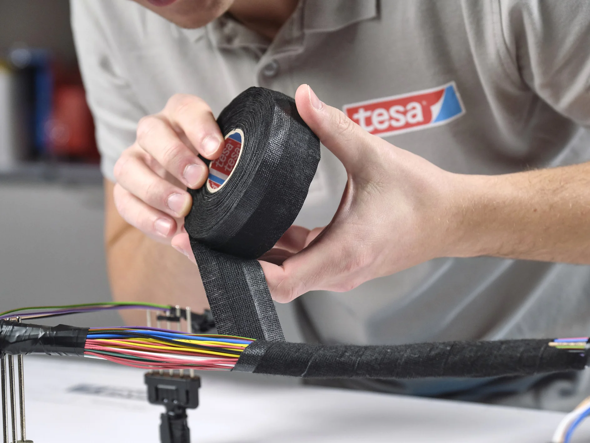 A person in a grey shirt is holding a roll of black tesa tape, wrapping it around a bundle of colorful cables on a table. The focus is on the hands and tesa tape, with a blurred background. (This text has been generated by AI)