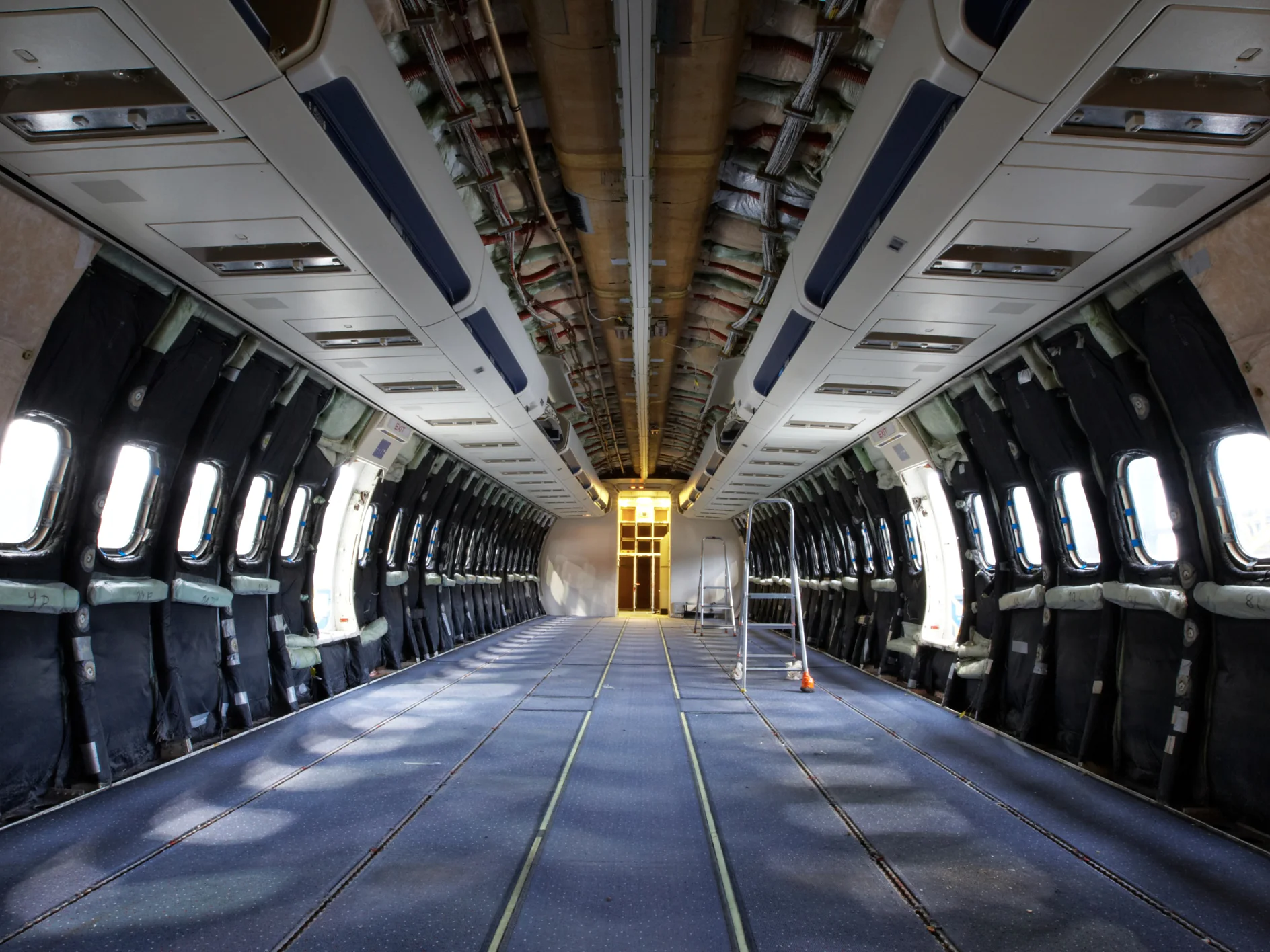 Interior view of an empty aircraft cabin under renovation. Seats and panels are removed, exposing insulation and wiring bound with tesa tape. A small ladder is placed near the center, and light enters through open windows along the sides. (This text has been generated by AI)