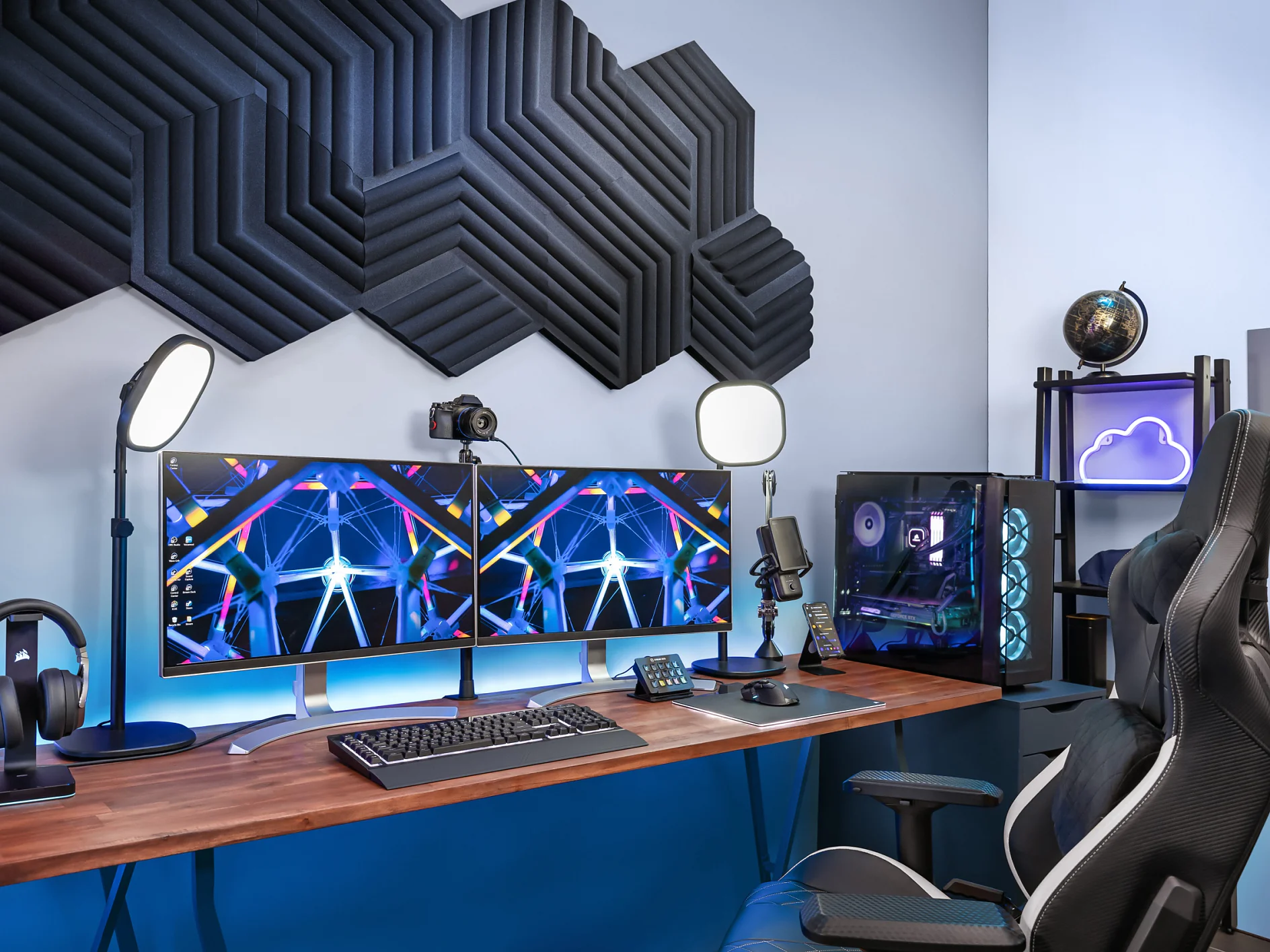 A modern gaming setup with dual monitors on a wooden desk, illuminated by LED lights. Gaming chair, keyboard, mouse, microphone, and headphones are visible. Acoustic panels are fastened to the wall using tesa tape, and a computer tower with RGB lighting is beside the desk. (This text has been generated by AI)