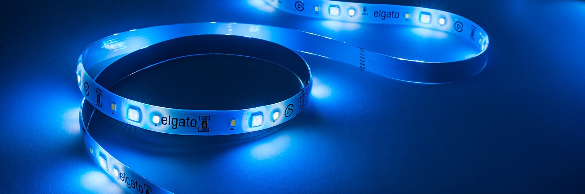 A flexible LED light strip emits a bright blue glow against a dark background. The strip is coiled in a loose, S-shaped pattern, showcasing individual LED components and tesa tape markings on the surface. (This text has been generated by AI)