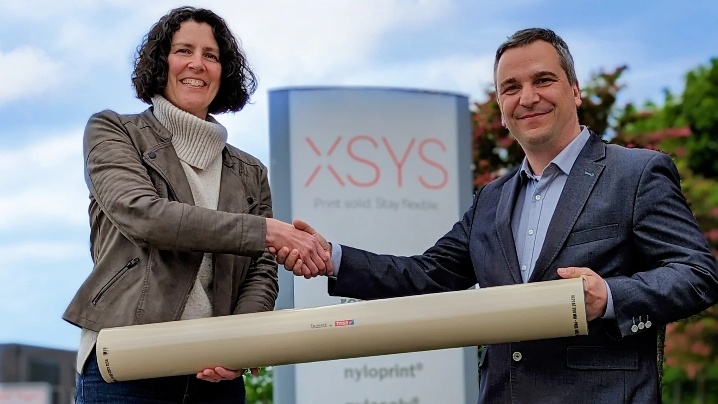 A woman and a man stand outside, shaking hands and holding a large cylindrical object. They are in front of a sign with the XSYS logo. The outdoor setting includes a cloudy sky and some greenery, with the product being securely held together using tesa tape. (This text has been generated by AI)