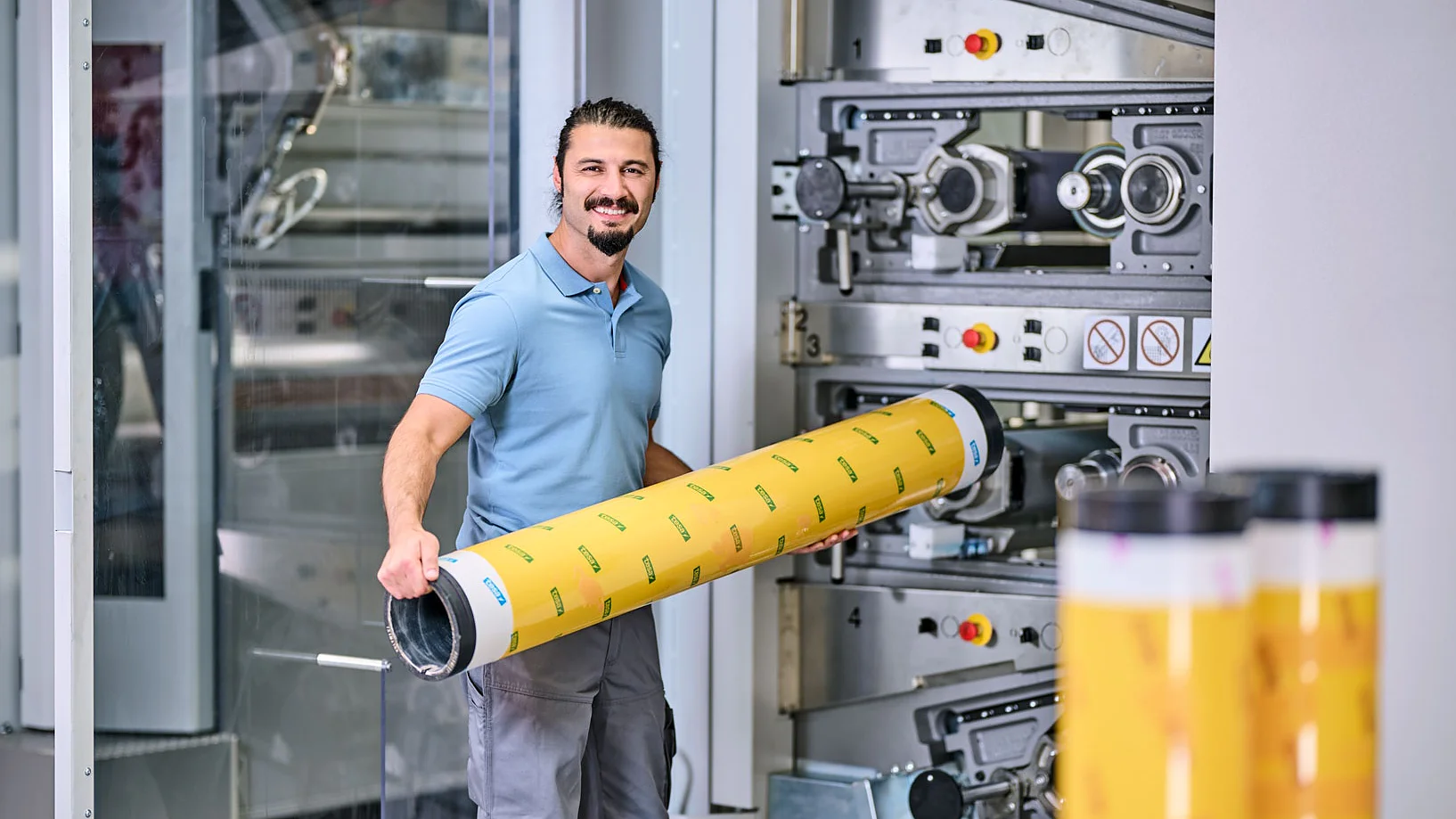 A man in a blue polo shirt is standing in front of industrial printing equipment, holding a large yellow cylindrical object with printed patterns. He is smiling, and other similar cylinders made with tesa tape are visible in the foreground. (This text has been generated by AI)