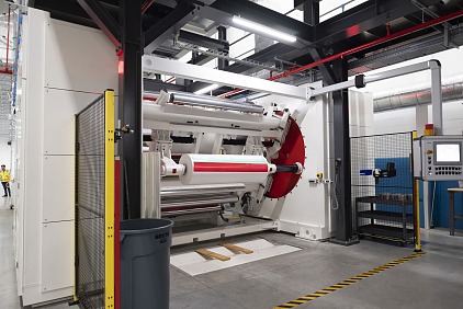 A large industrial machine with rollers and a red component stands in a factory setting. A control panel is visible on the right, and tesa tape marks the area on the floor. The ceiling has visible pipes and ducts.