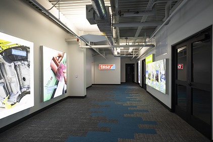 A corridor with illuminated displays on either side showcases automotive and other products. The walls are gray, and the ceiling has exposed ducts. A sign with the logo tesa tape is visible at the end of the corridor. The carpet has blue accents.