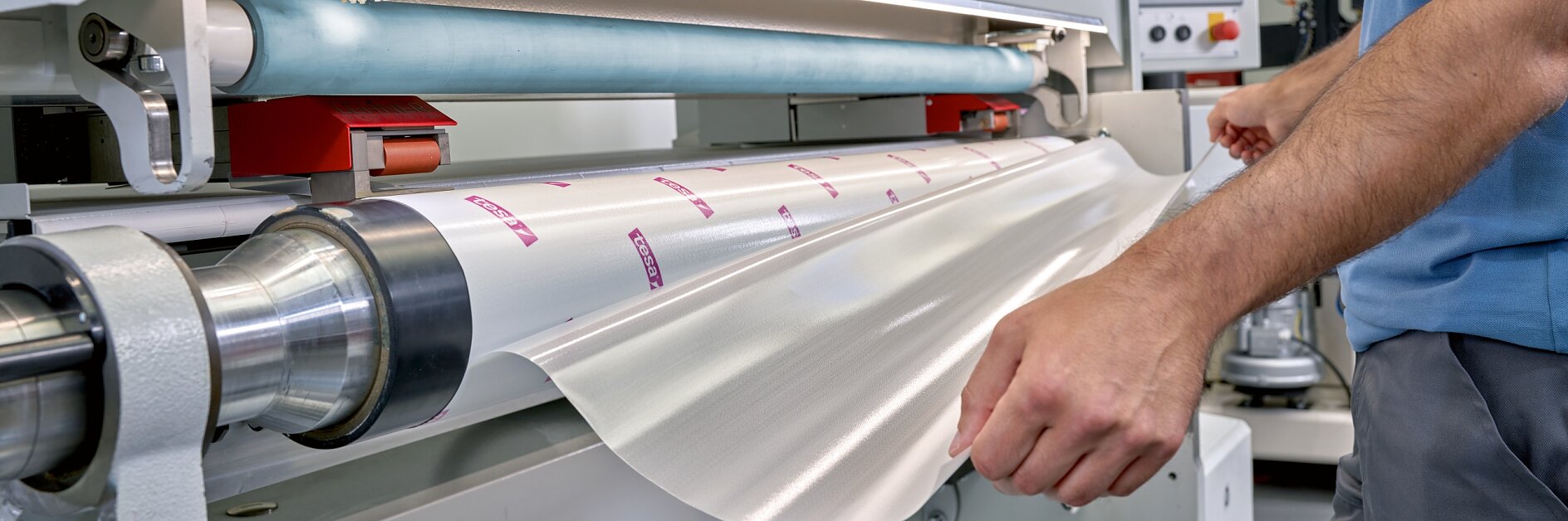 A person operates a large industrial machine, feeding a roll of clear plastic material secured with tesa tape into it. The machine has a control panel with display and buttons. The person, wearing a blue shirt, is adjusting the material as it enters the machine. (This text has been generated by AI)