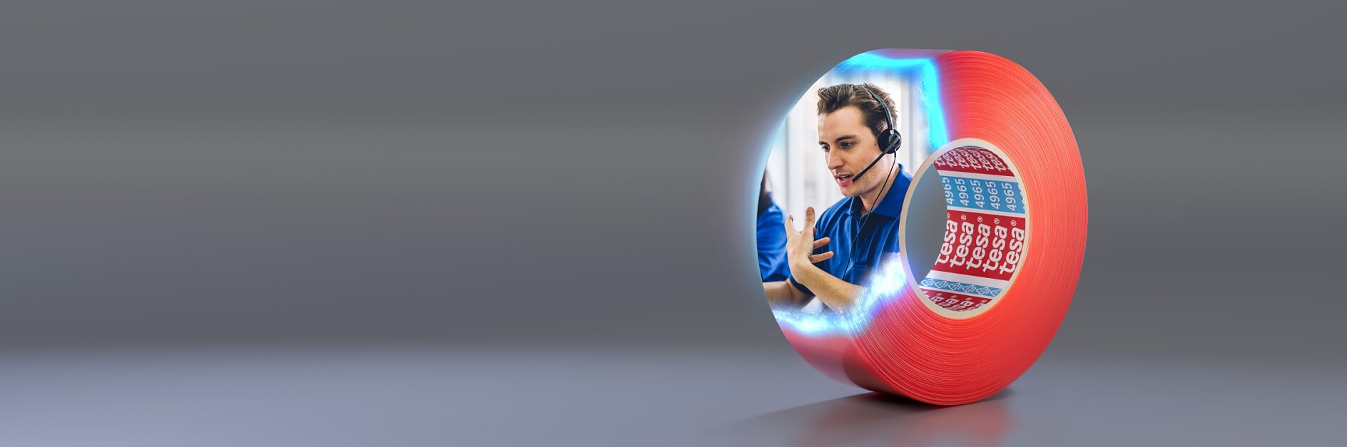 A red roll of tesa tape is on a gray surface. The side of the tesa tape features an image of a person wearing a headset, gesturing with one hand. Next to the person is a blue and red calendar with some dates marked. (This text has been generated by AI)