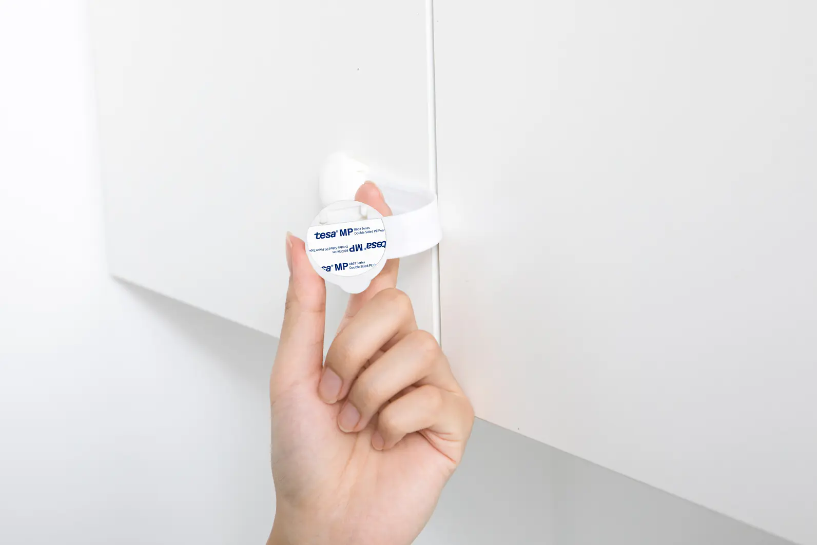 A hand is seen holding a white plastic latch on a cabinet door. The latch has blue text on it and is used to secure the door, likely as a child safety feature. The cabinet appears to be white and part of a neatly painted surface, with tesa tape used in its assembly or installation process. (This text has been generated by AI)