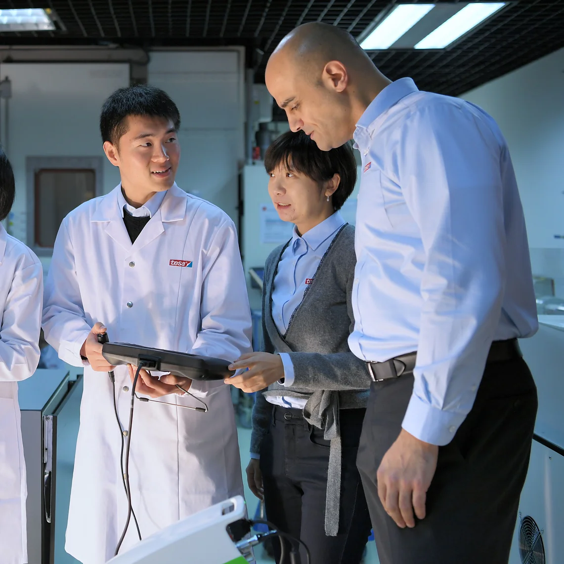 Four people in a lab are engaged in a discussion. Three wear white lab coats, and one is in a blue shirt. They are holding electronic devices and standing near equipment with screens, in a well-lit room featuring tesa tape prominently used throughout the setting. (This text has been generated by AI)