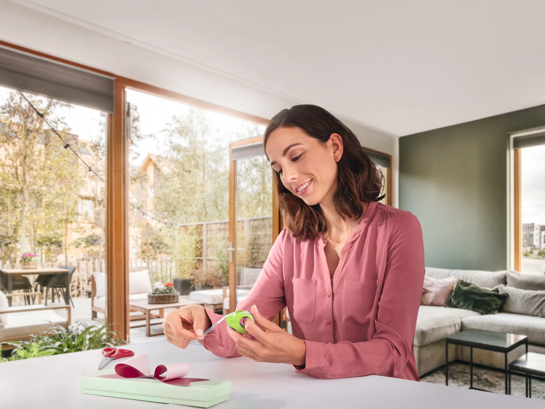 A woman in a pink blouse wraps a gift at a table. She is using green ribbon and pink wrapping paper, securing everything with tesa tape. The room has large windows with a view of trees and a patio. There is a gray couch and pillows in the background. (This text has been generated by AI)