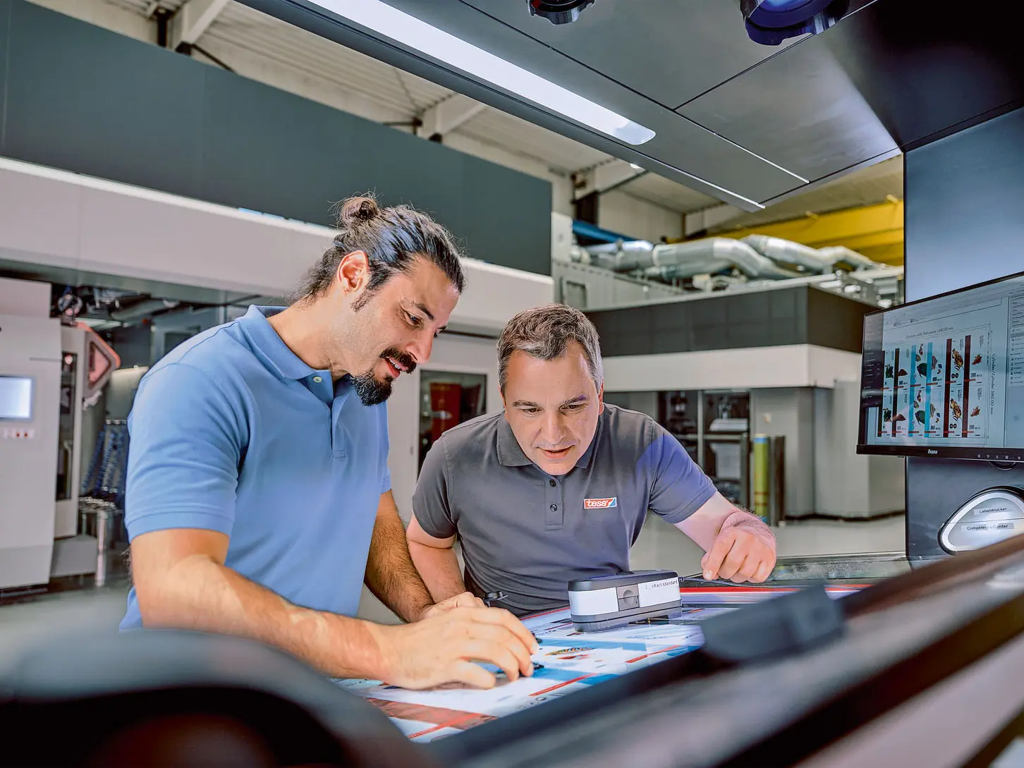 Two men are examining printed pages under a light in an industrial setting. One holds a magnifying glass, while the other points at the paper secured with tesa tape. A computer monitor displaying graphics is visible on the side. Both are wearing casual shirts. (This text has been generated by AI)
