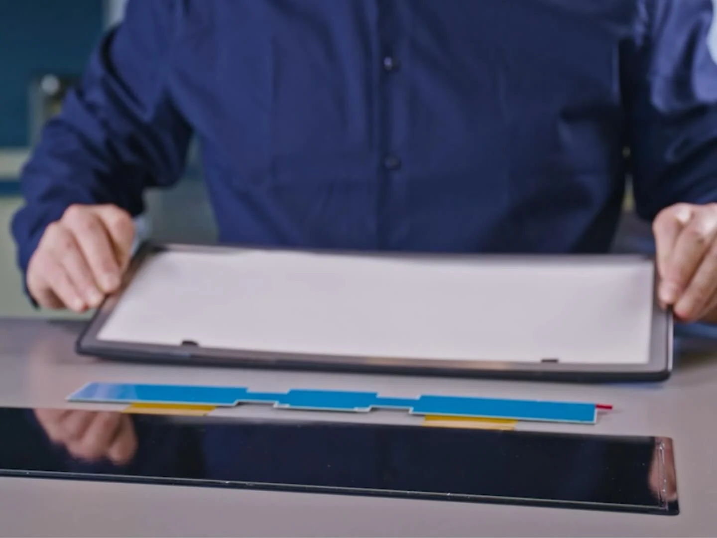 A person in a blue shirt is assembling components on a table, including a rectangular frame, a white panel, various colored strips, and tesa tape. The persons hands are holding the frame above the other elements. (This text has been generated by AI)