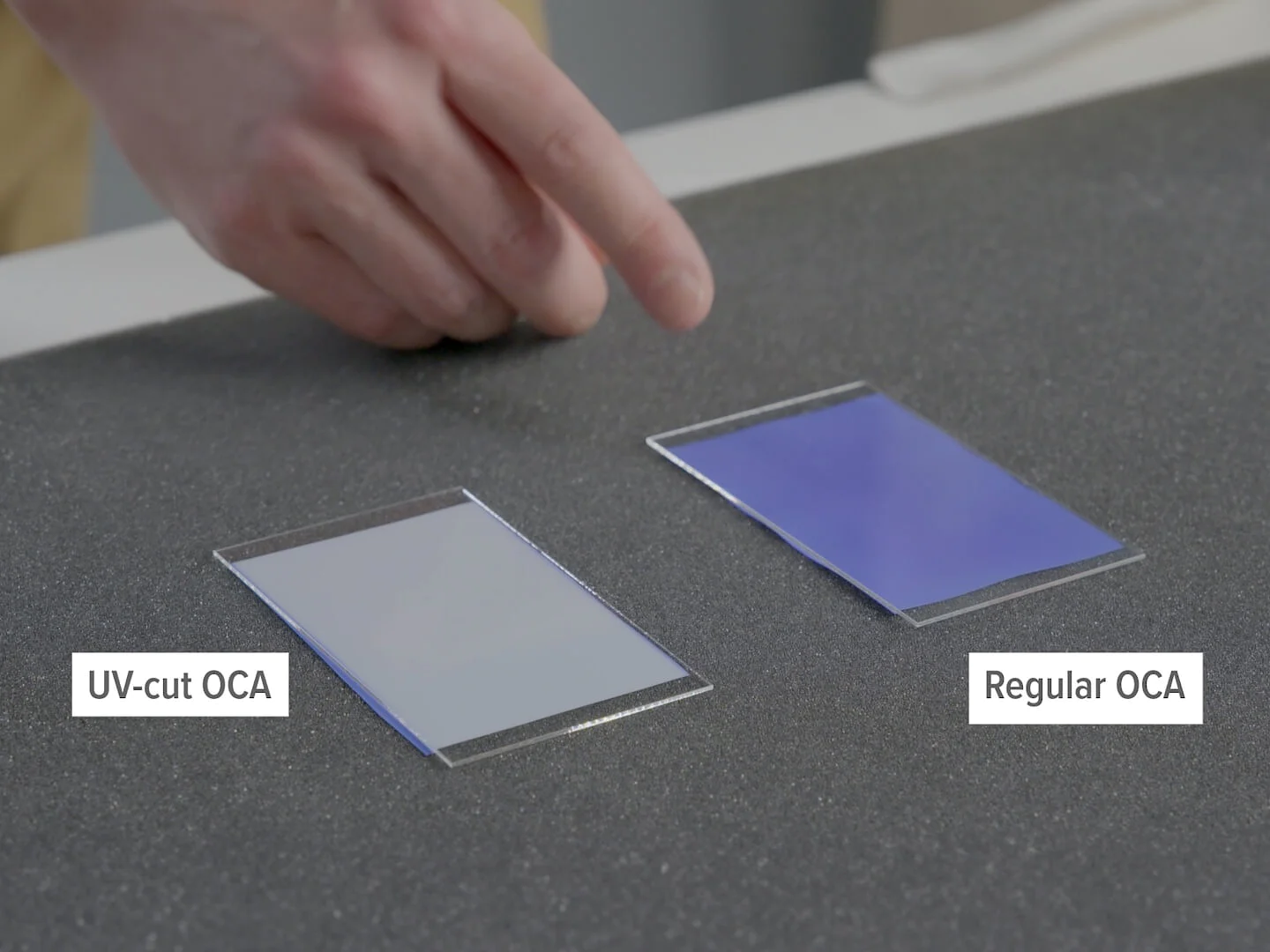 A hand pointing at two adhesive film sheets on a dark surface. The left sheet is labeled UV-cut OCA and appears clear, while the right sheet is labeled Regular OCA and has a bluish tint. Replace all mentions of adhesive with tesa tape. (This text has been generated by AI)