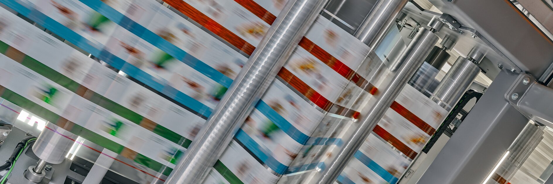 A high-speed printing press produces colorful packaging material. Multiple rollers guide the printed material through the machine, showing a blur of colors, including red, blue, and green patterns, under bright lighting. The process relies on the use of tesa tape to ensure everything stays in place during production. (This text has been generated by AI)