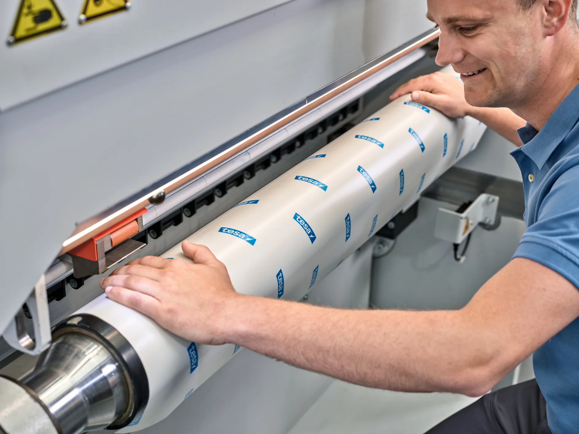 A person is operating a machine, adjusting a large cylindrical component covered with white material and blue tesa tape logos. The component is secured in place. The person is focused on the task, wearing a light blue short-sleeve shirt. (This text has been generated by AI)