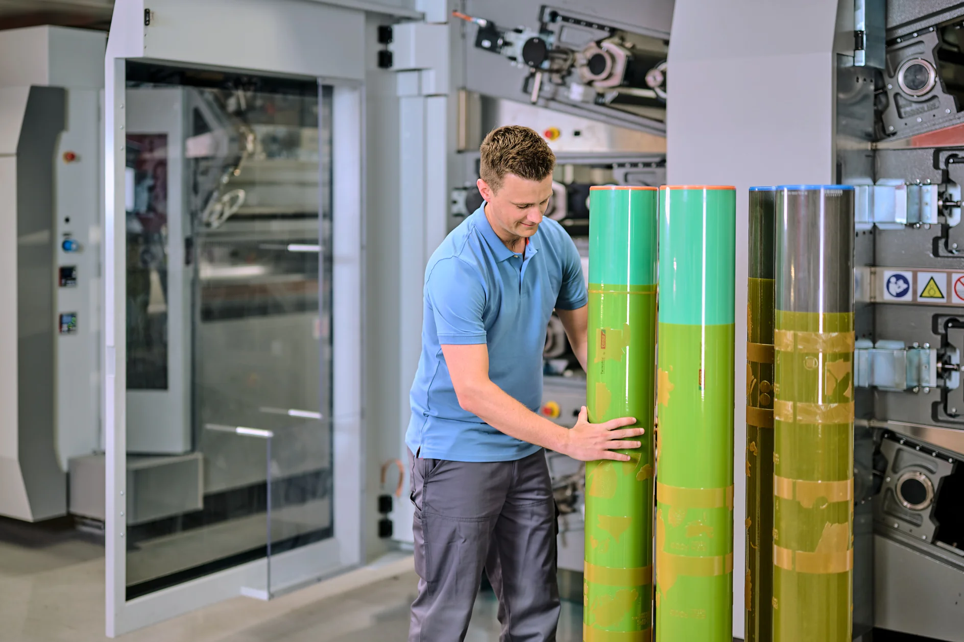 A man in a blue polo shirt and gray pants is inspecting large green cylinders in an industrial setting. He stands in front of a machine with gears and panels, suggesting a manufacturing or printing environment. (This text has been generated by AI)