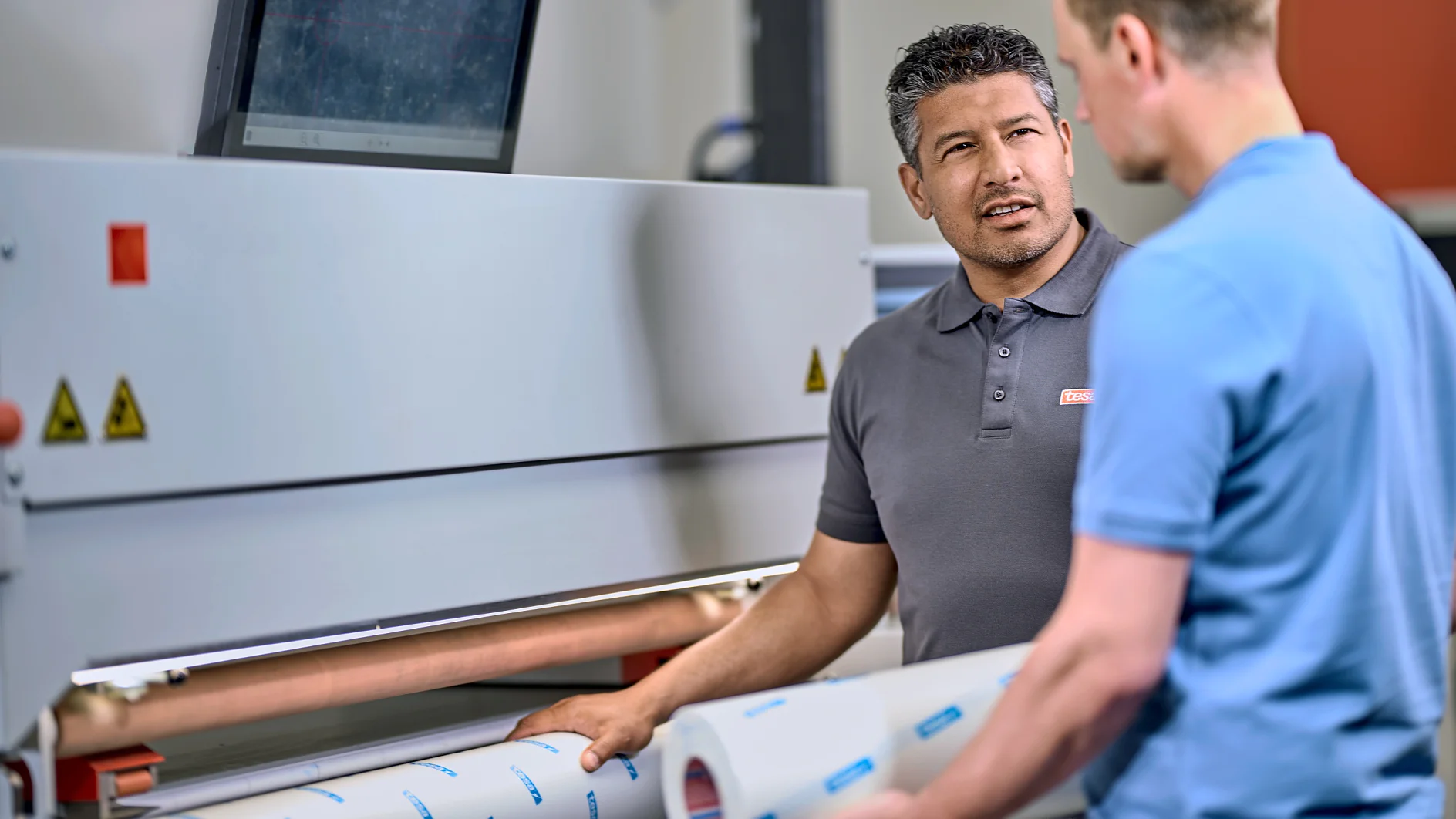 Two men are standing in front of a large industrial machine, discussing something. One man is holding a roll of tesa tape. A computer monitor is mounted on top of the machine. They are in a well-lit workspace. (This text has been generated by AI)
