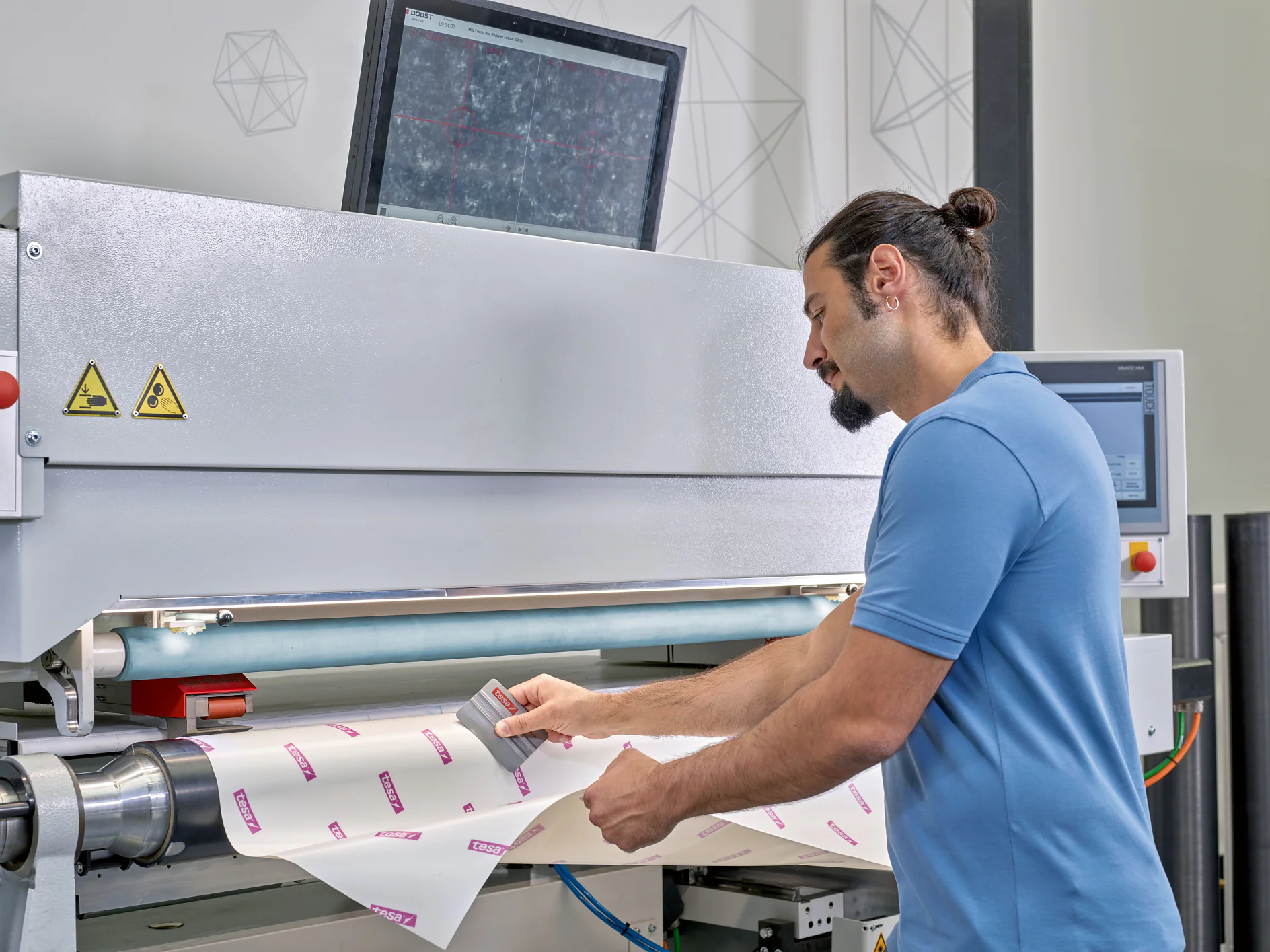A man in a blue shirt operates a machine, adjusting a piece of paper or material with printed text. A monitor is mounted above the machine, displaying a detailed screen. Control panels are visible, with geometric patterns on the wall. In his hand, he carefully applies tesa tape to secure the material in place, ensuring precision and stability throughout the process. (This text has been generated by AI)