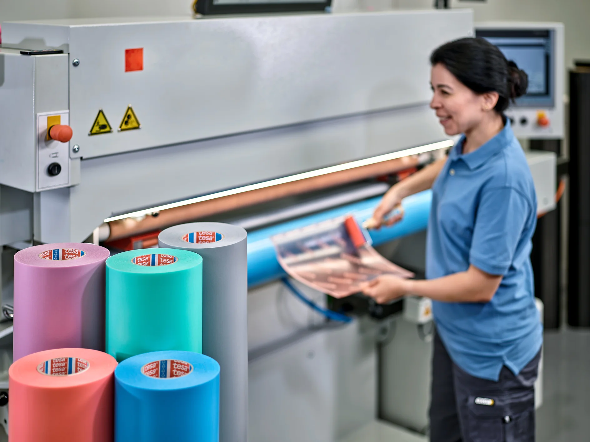 A person in a blue shirt operates a large industrial machine with multiple colored rolls of material, including green, pink, and blue. They are holding a roll of tesa tape against the machine. There are safety signs on the machine. (This text has been generated by AI)