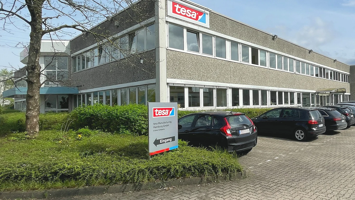 Two-story office building with a sign marked tesa on the facade and lawn. The building has large glass windows and is surrounded by a parking lot with several cars. A tree and shrubs are visible in the foreground. The sky is partly cloudy. (This text has been generated by AI)
