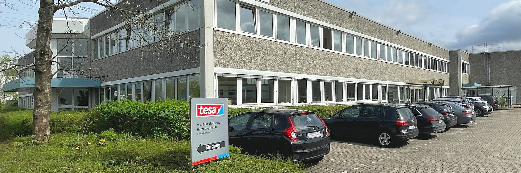 Two-story office building with a sign marked tesa on the facade and lawn. The building has large glass windows and is surrounded by a parking lot with several cars. A tree and shrubs are visible in the foreground. The sky is partly cloudy. (This text has been generated by AI)