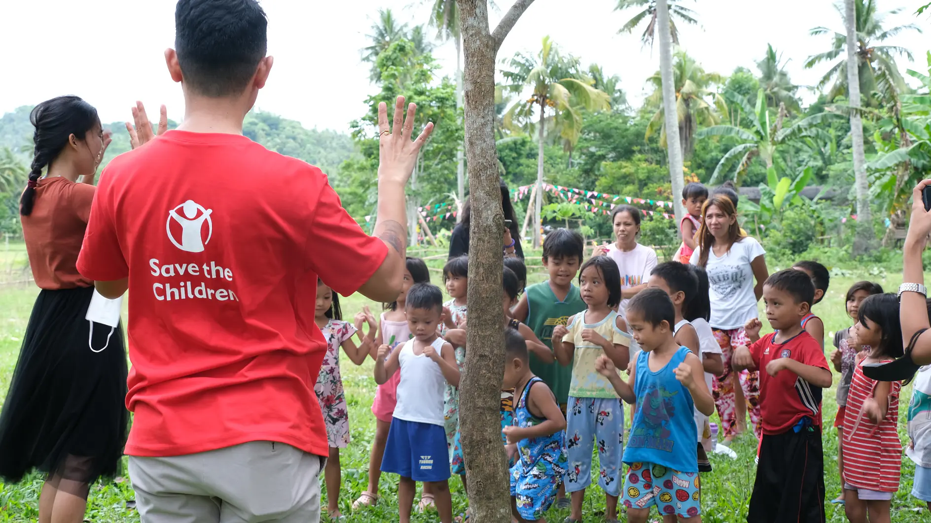 A man in a red Save the Children shirt stands facing a group of children outdoors. The children, wearing casual clothing, are gathered around him. A woman is also interacting with the children. The setting is a grassy area with trees and people in the background. (This text has been generated by AI)