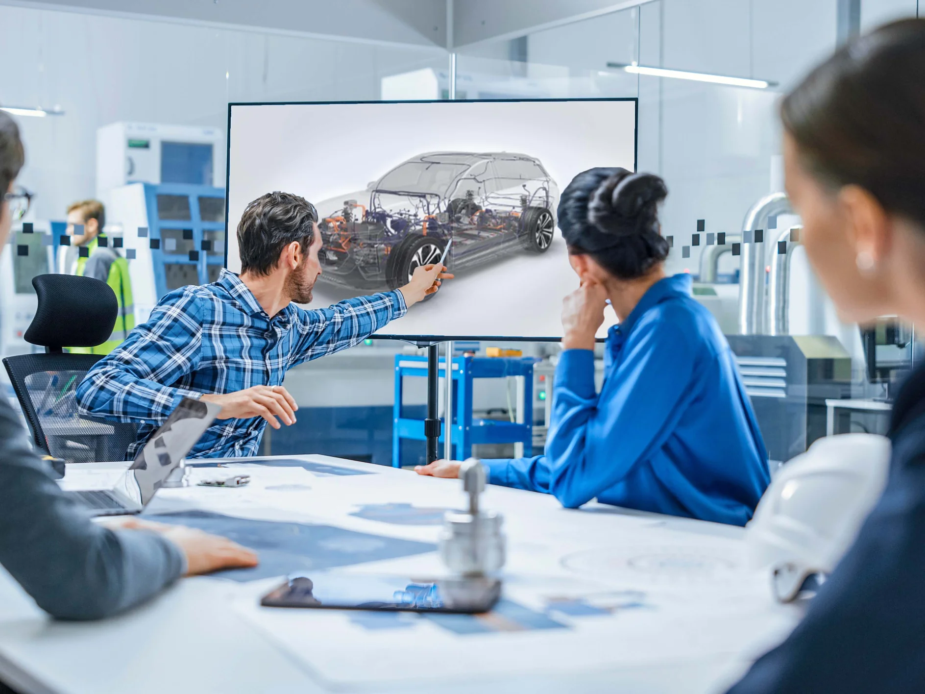 A group of four people in a modern office setting look at a screen displaying a cars internal components. A man in a plaid shirt points at the screen. Various technical documents, equipment, and tesa tape are on the table. (This text has been generated by AI)
