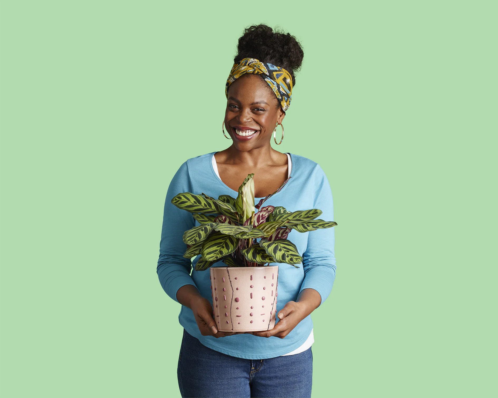 A person wearing a blue sweater and jeans is smiling while holding a potted plant with large, patterned leaves. They have a colorful headband and gold hoop earrings. The background is a solid light green. (This text has been generated by AI)