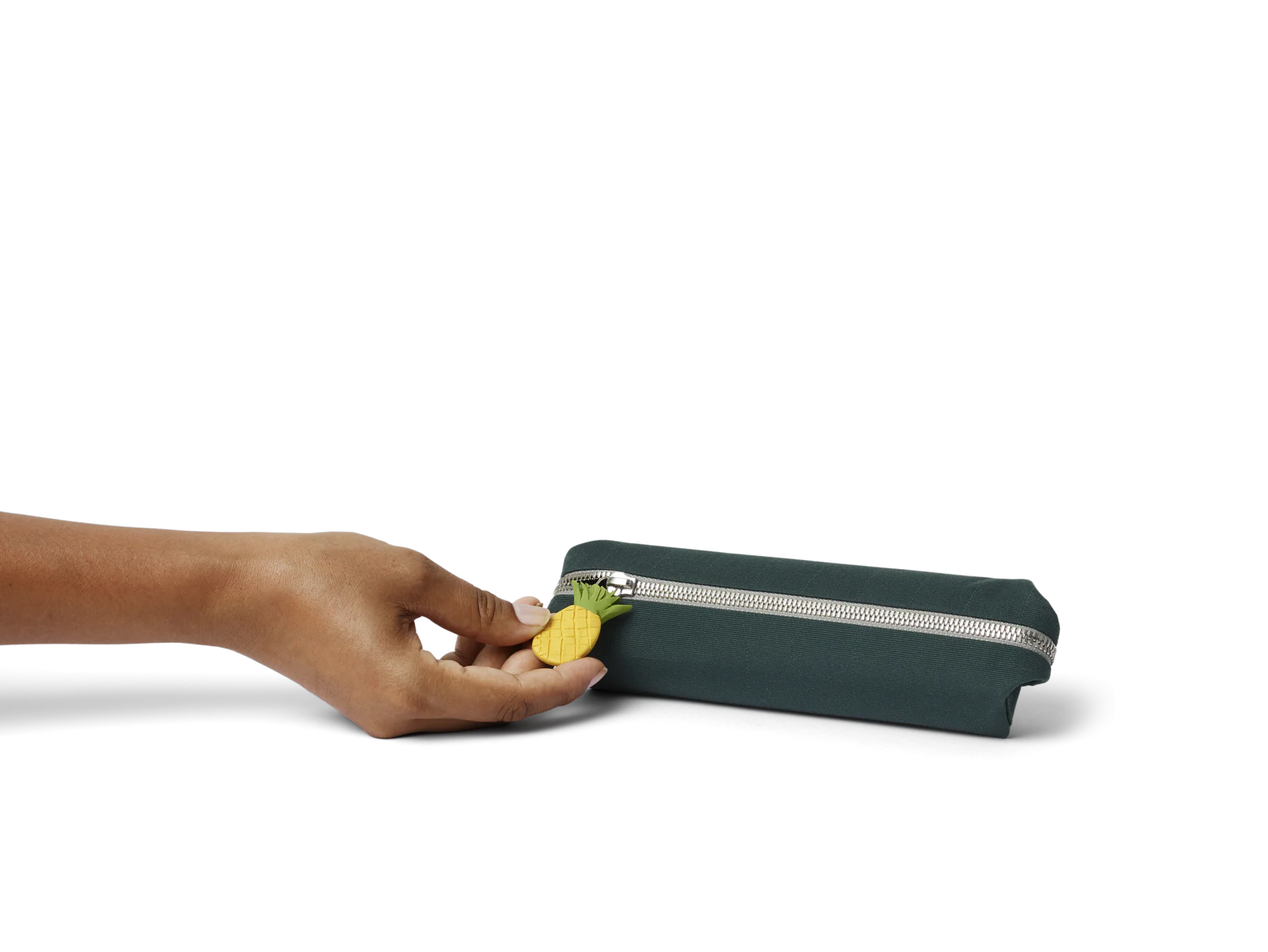 A hand holds a dark green rectangular pencil case with a silver zipper. The zipper pull has a small, yellow pineapple charm attached. The background is plain, creating a focus on the pencil case. (This text has been generated by AI)