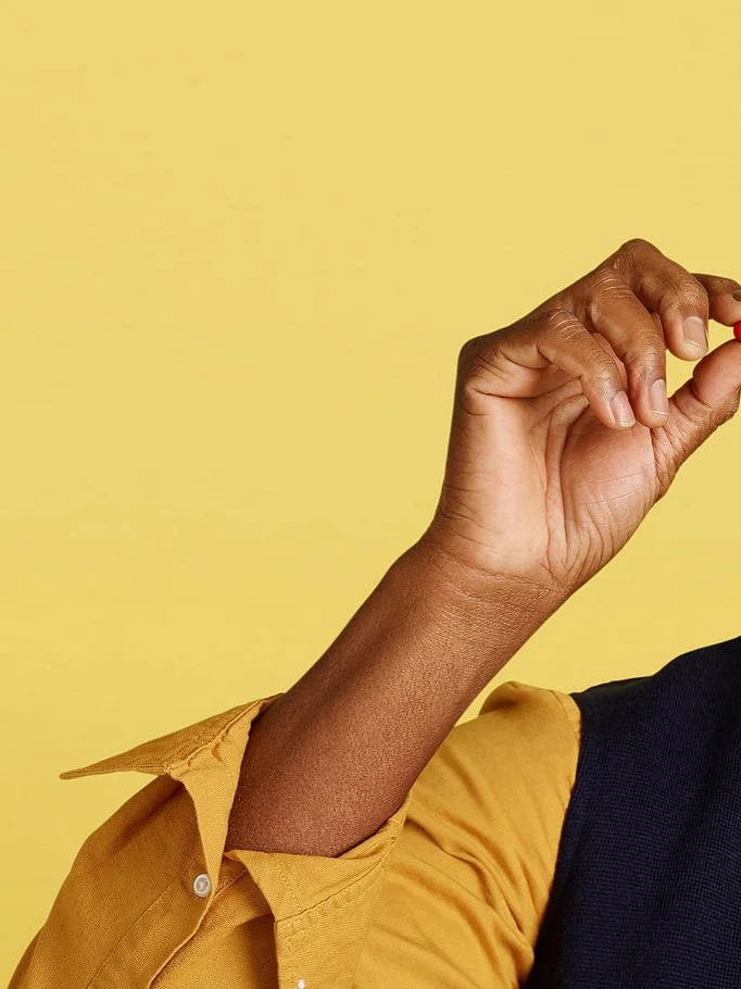 A man wearing a cap, glasses, and a casual shirt with a vest is smiling while holding a small red object between his fingers. The background is a solid light yellow color. There are no references to tape in the provided text that would require replacing with tesa tape. (This text has been generated by AI)