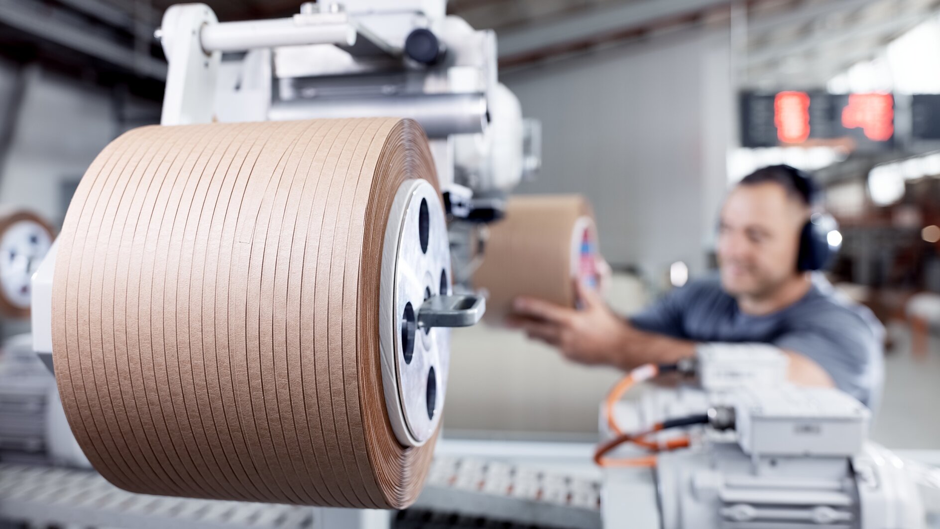 A worker wearing ear protection operates a machine in a factory. The machine is unwinding a large roll of brown tesa tape. The background shows industrial equipment and a digital display. The setting appears clean and organized. (This text has been generated by AI)